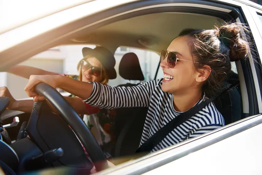 Two attractive young girlfriends wearing sunglasses talking and laughing together while driving in a car through the city on a sunny day