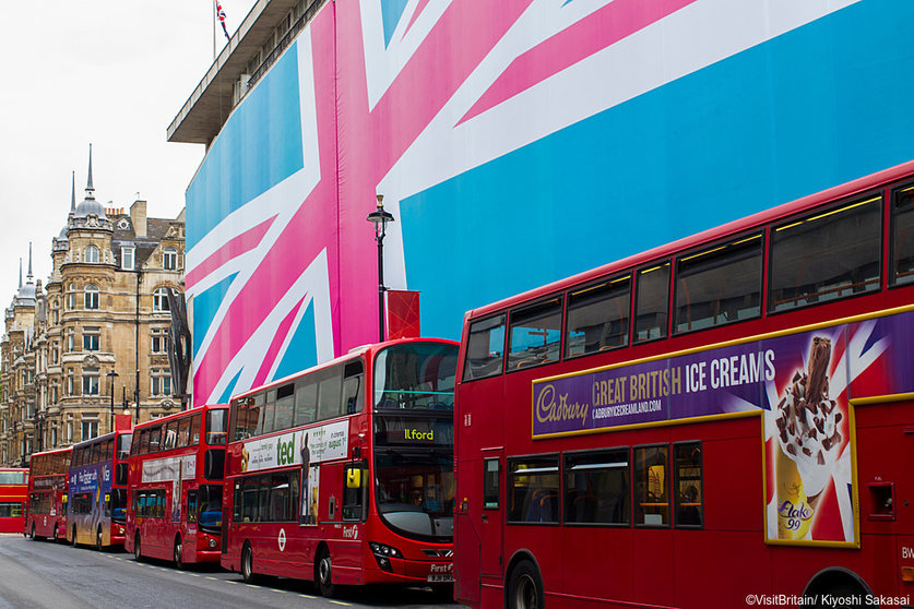 A row of red double decker buses on a London street. A building with the exterior decorated in the colours of the Union Jack flag.