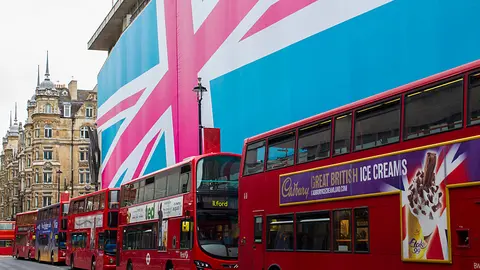 A row of red double decker buses on a London street. A building with the exterior decorated in the colours of the Union Jack flag.